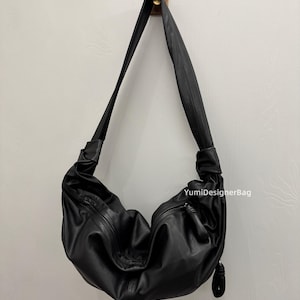 May include: Black leather shoulder bag with a curved design. The bag has a long strap and a zipper closure. The bag is hanging on a wooden hook against a white wall. The text "YumiDesignerBag" is visible on the bag.