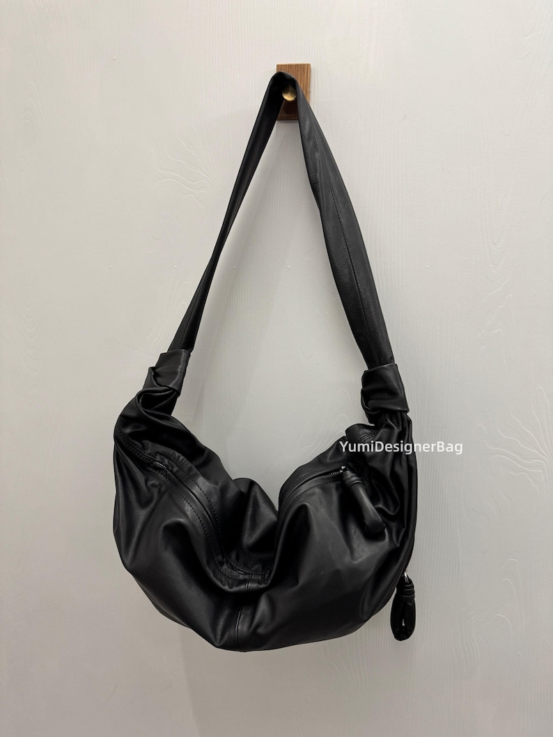 May include: Black leather shoulder bag with a curved design. The bag has a long strap and a zipper closure. The bag is hanging on a wooden hook against a white wall. The text "YumiDesignerBag" is visible on the bag.