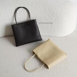 May include: Two square-shaped tote bags with thin shoulder straps, one in solid black and one in solid beige. The black bag is positioned upright, while the beige bag lies flat. The text YumiDesignerBag is printed in black near the black bag.