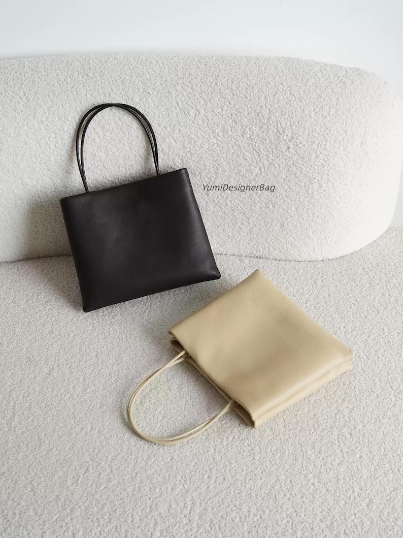 May include: Two square-shaped tote bags with thin shoulder straps, one in solid black and one in solid beige. The black bag is positioned upright, while the beige bag lies flat. The text YumiDesignerBag is printed in black near the black bag.