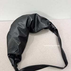 May include: A black, crescent-shaped shoulder bag made of a smooth, leather-like material. The bag features a knotted handle and a curved design. The text "YumiDesignerBag" is visible on the right side of the image.