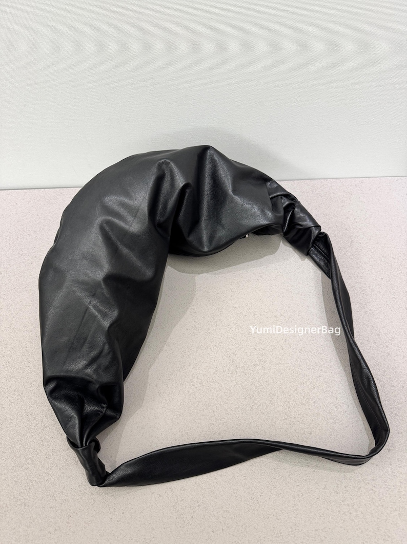 May include: A black, crescent-shaped shoulder bag made of a smooth, leather-like material. The bag features a knotted handle and a curved design. The text "YumiDesignerBag" is visible on the right side of the image.