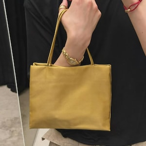 May include: A rectangular mustard yellow handbag with a smooth, matte finish and two thin, matching shoulder straps. The bag features a clean, minimalist design with a structured silhouette and a simple, unadorned front panel, providing a compact storage solution.