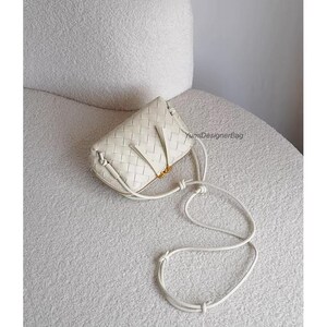 K&ouml;nnte beinhalten: Eine cremefarbene Crossbody-Tasche aus geflochtenem Leder mit einem langen, verstellbaren Riemen. Die Tasche hat eine rechteckige Form mit Rei&szlig;verschluss und goldfarbenen Beschl&auml;gen. Die Tasche liegt auf einer wei&szlig;en, strukturierten Oberfl&auml;che.