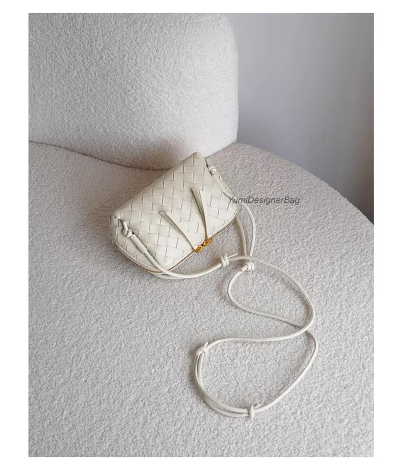 K&ouml;nnte beinhalten: Eine cremefarbene Crossbody-Tasche aus geflochtenem Leder mit einem langen, verstellbaren Riemen. Die Tasche hat eine rechteckige Form mit Rei&szlig;verschluss und goldfarbenen Beschl&auml;gen. Die Tasche liegt auf einer wei&szlig;en, strukturierten Oberfl&auml;che.