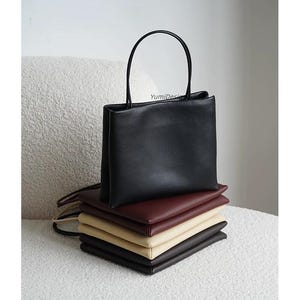May include: A stack of five rectangular handbags in black, burgundy, beige, and dark brown. The top black bag stands upright with a thin handle, while the others are folded flat. The text YumiDesign is printed in small black letters on the top bag.