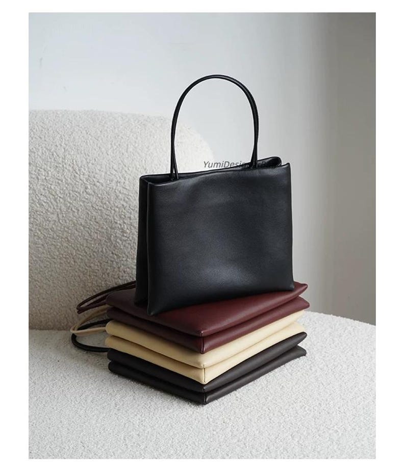 May include: A stack of five rectangular handbags in black, burgundy, beige, and dark brown. The top black bag stands upright with a thin handle, while the others are folded flat. The text YumiDesign is printed in small black letters on the top bag.