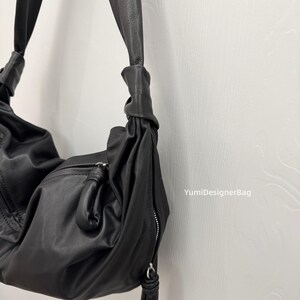 May include: A black leather shoulder bag with a knotted strap design. The bag features a zippered closure and a soft, slouchy shape. The bag is hanging against a white wall.