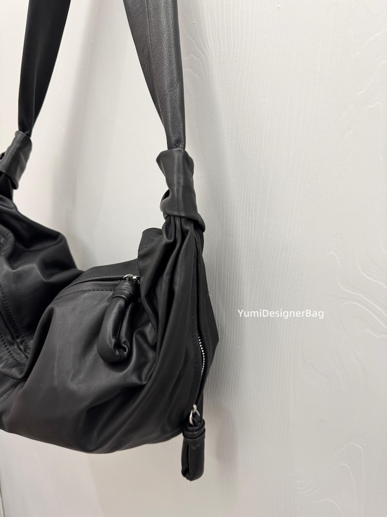 May include: A black leather shoulder bag with a knotted strap design. The bag features a zippered closure and a soft, slouchy shape. The bag is hanging against a white wall.