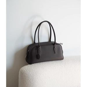 May include: A dark brown handbag with two curved handles. The bag has a rounded shape with a zipper closure and a small tag attached. The bag is sitting on a white, textured surface against a white wall.