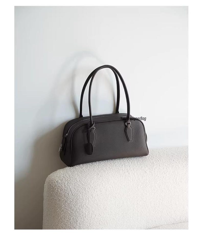May include: A dark brown handbag with two curved handles. The bag has a rounded shape with a zipper closure and a small tag attached. The bag is sitting on a white, textured surface against a white wall.