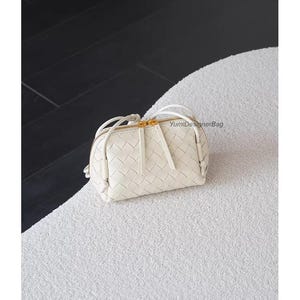 K&ouml;nnte beinhalten: Eine kleine, cremefarbene Handtasche aus geflochtenem Leder mit goldenem Rei&szlig;verschluss und Quastendetail. Die Tasche hat eine runde Form und ein gewebtes Muster. Die Tasche liegt auf einer wei&szlig;en, strukturierten Oberfl&auml;che.