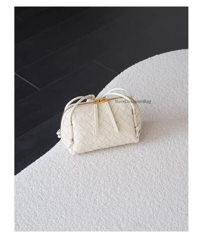 K&ouml;nnte beinhalten: Eine kleine, cremefarbene Handtasche aus geflochtenem Leder mit goldenem Rei&szlig;verschluss und Quastendetail. Die Tasche hat eine runde Form und ein gewebtes Muster. Die Tasche liegt auf einer wei&szlig;en, strukturierten Oberfl&auml;che.