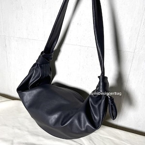 May include: Black shoulder bag with a knotted strap design. The bag has a crescent shape and is made of a smooth, matte material. The strap is long and adjustable, allowing it to be worn over the shoulder or across the body. The bag is a solid black color.