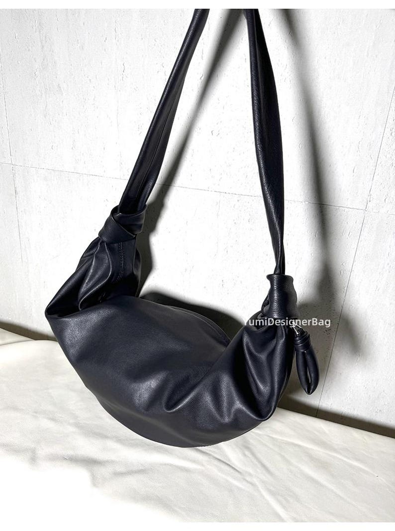 May include: Black shoulder bag with a knotted strap design. The bag has a crescent shape and is made of a smooth, matte material. The strap is long and adjustable, allowing it to be worn over the shoulder or across the body. The bag is a solid black color.