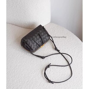 K&ouml;nnte beinhalten: Schwarze Crossbody-Tasche aus geflochtenem Leder mit goldenem Rei&szlig;verschluss und Riemen. Die Tasche hat eine rechteckige Form mit abgerundeten Kanten. Der Riemen ist d&uuml;nn und verstellbar. Die Tasche liegt auf einer wei&szlig;en Oberfl&auml;che.