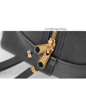 May include: Close-up of a black leather bag with a gold zipper and matching gold zipper pulls. The bag has a strap and the text "YumiDesignerBag" is visible.