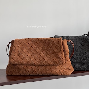 Könnte beinhalten: Eine braune Umhängetasche aus Wildleder mit Flechtmuster und Umschlagklappe sowie dünnen Seitenriemen. Der Text YumiDesignerBag ist über der Tasche zu sehen. Eine zweite schwarze geflochtene Tasche ist rechts neben der braunen Tasche zu sehen.
