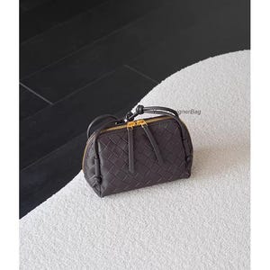 K&ouml;nnte beinhalten: Eine dunkelbraune, gewebte Lederhandtasche mit goldenem Rei&szlig;verschluss und kleinem Griff. Die Tasche ist oval und liegt auf einer wei&szlig;en, strukturierten Oberfl&auml;che. Die Tasche hat einen Rei&szlig;verschluss und einen kleinen Griff.
