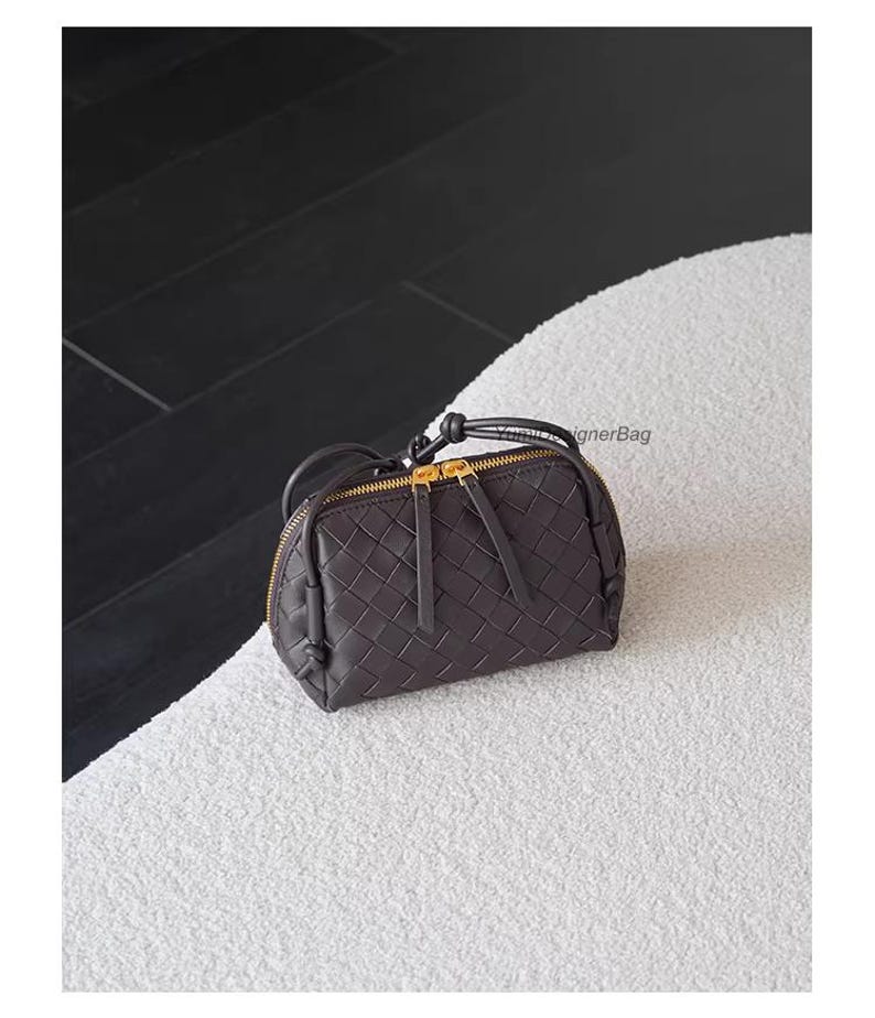 K&ouml;nnte beinhalten: Eine dunkelbraune, gewebte Lederhandtasche mit goldenem Rei&szlig;verschluss und kleinem Griff. Die Tasche ist oval und liegt auf einer wei&szlig;en, strukturierten Oberfl&auml;che. Die Tasche hat einen Rei&szlig;verschluss und einen kleinen Griff.