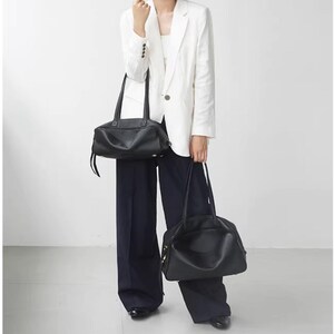 May include: Two black leather handbags with long handles. One is rectangular, the other is rounded. The bags are held by a person wearing a white blazer and dark blue pants. The background is plain white.