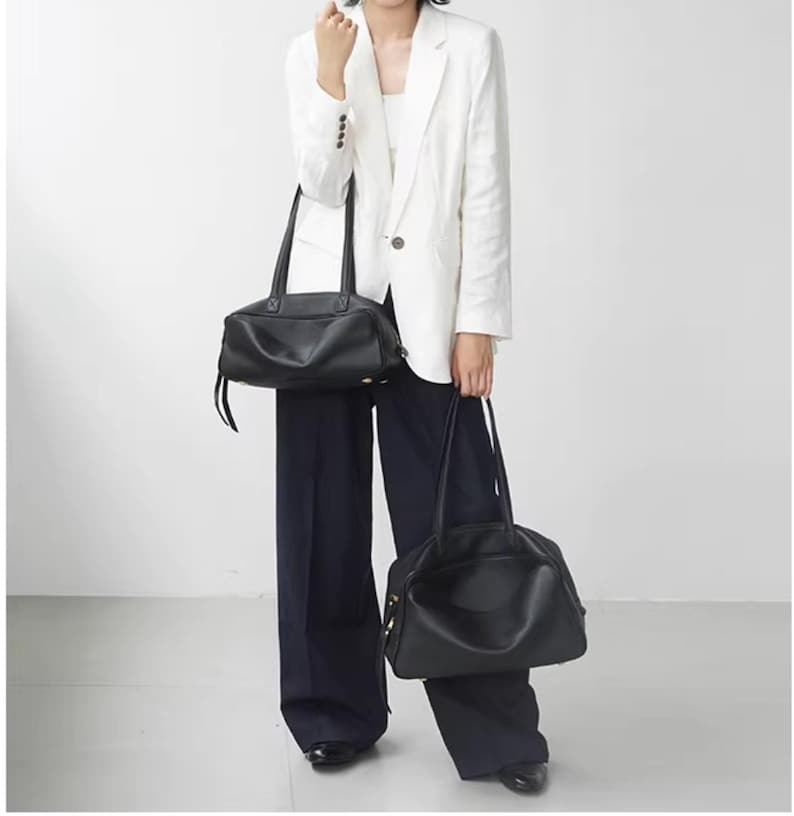 May include: Two black leather handbags with long handles. One is rectangular, the other is rounded. The bags are held by a person wearing a white blazer and dark blue pants. The background is plain white.