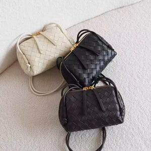 K&ouml;nnte beinhalten: Drei kleine, gewebte Lederhandtaschen in Creme, Schwarz und Dunkelbraun. Jede Tasche hat einen Rei&szlig;verschluss mit goldfarbenem Rei&szlig;verschluss und einen passenden Riemen. Die Taschen liegen auf einer weichen, strukturierten Oberfl&auml;che.