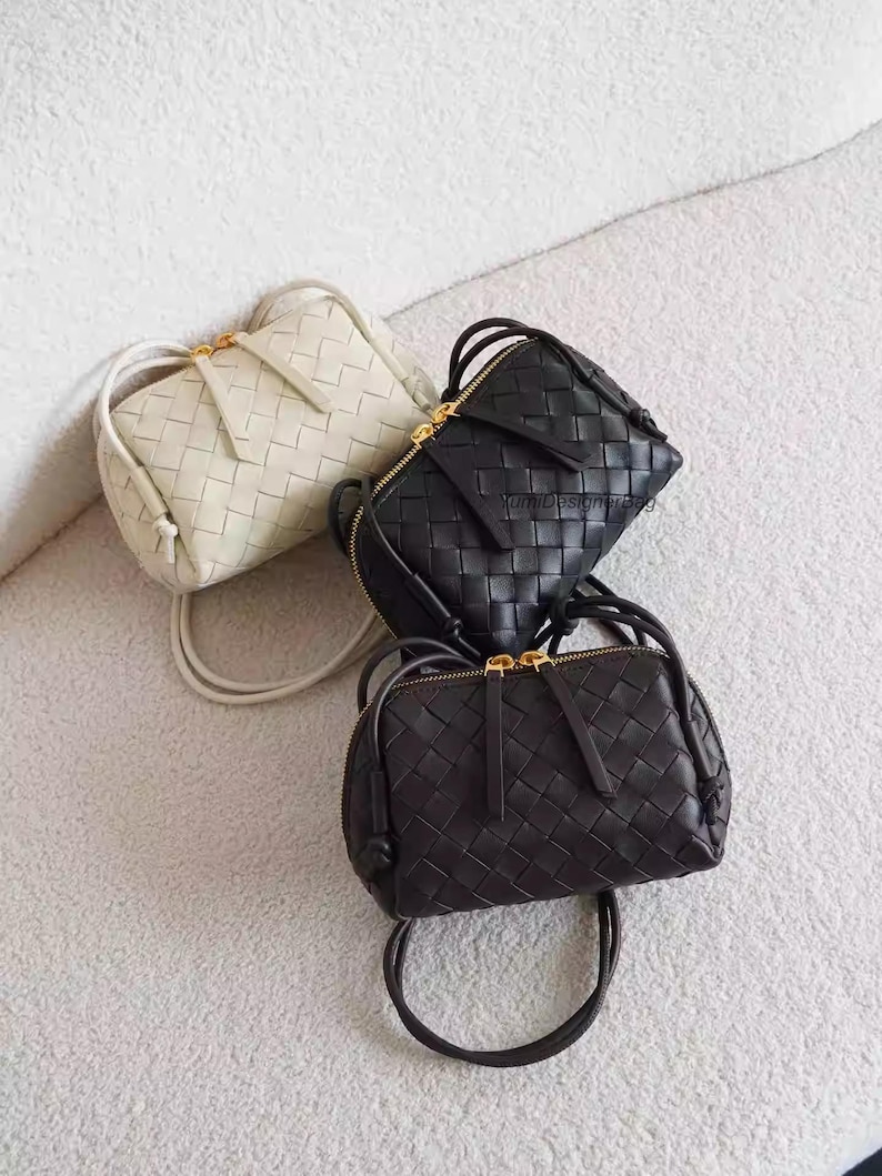 K&ouml;nnte beinhalten: Drei kleine, gewebte Lederhandtaschen in Creme, Schwarz und Dunkelbraun. Jede Tasche hat einen Rei&szlig;verschluss mit goldfarbenem Rei&szlig;verschluss und einen passenden Riemen. Die Taschen liegen auf einer weichen, strukturierten Oberfl&auml;che.