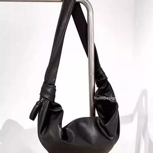 May include: Black leather shoulder bag with a knotted strap. The bag has a crescent shape and a zipper closure. The bag is hanging on a metal bar. The text "YumiDesign" is visible on the bag.