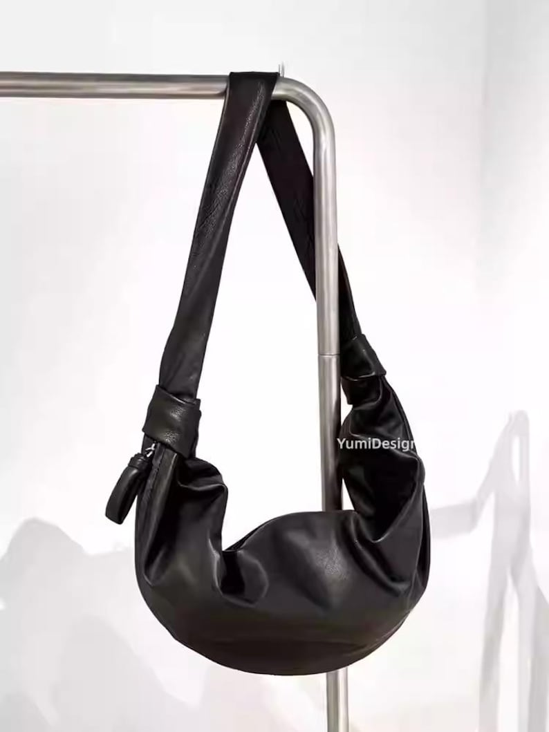 May include: Black leather shoulder bag with a knotted strap. The bag has a crescent shape and a zipper closure. The bag is hanging on a metal bar. The text "YumiDesign" is visible on the bag.