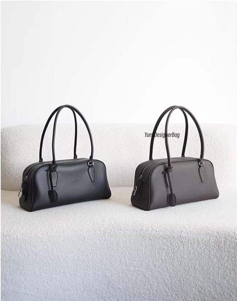 May include: Two leather handbags, one black and one dark gray, with matching handles and zipper closures. Each bag has a rectangular shape and a small tag attached to the zipper. The bags are displayed on a white, textured surface.