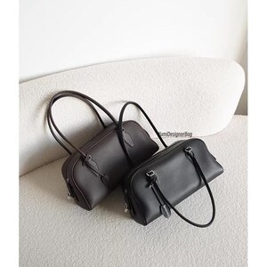 May include: Two leather handbags. One is dark brown, the other is black. Both have a rectangular shape with rounded corners, top handles, and a small strap detail. The bags are displayed on a light-colored, textured surface.