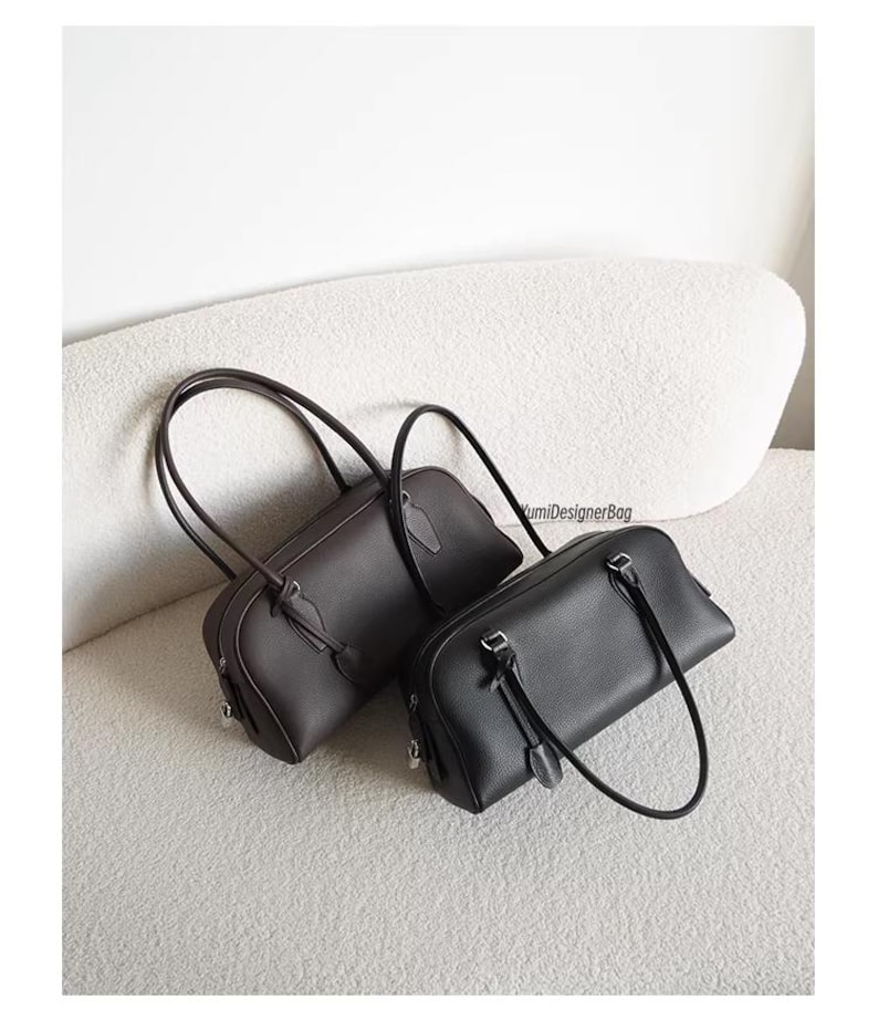May include: Two leather handbags. One is dark brown, the other is black. Both have a rectangular shape with rounded corners, top handles, and a small strap detail. The bags are displayed on a light-colored, textured surface.