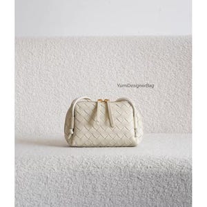 K&ouml;nnte beinhalten: Cremefarbene, gewebte Lederhandtasche mit rechteckiger Form und Rei&szlig;verschluss oben. Die Tasche hat einen goldfarbenen Rei&szlig;verschluss und einen kleinen Griff. Der Text "YumiDesignerBag" ist sichtbar.