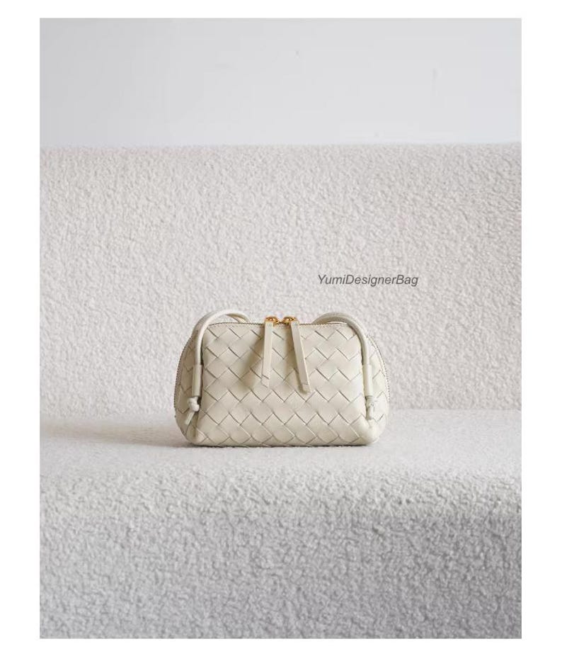 K&ouml;nnte beinhalten: Cremefarbene, gewebte Lederhandtasche mit rechteckiger Form und Rei&szlig;verschluss oben. Die Tasche hat einen goldfarbenen Rei&szlig;verschluss und einen kleinen Griff. Der Text "YumiDesignerBag" ist sichtbar.