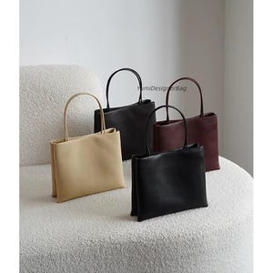 May include: Four rectangular handbags with thin top handles, featuring the text YumiDesignerBag. The bags are available in beige, black, and dark red, each with a smooth finish and a structured, upright design with a flat base and clean lines.