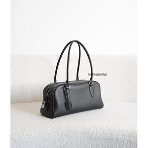 May include: Black leather handbag with a rectangular shape and two curved handles. The bag features a zipper closure, a small lock, and a decorative leather accent. The bag is sitting on a white, textured surface.