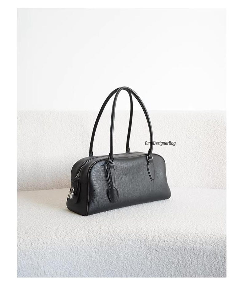May include: Black leather handbag with a rectangular shape and two curved handles. The bag features a zipper closure, a small lock, and a decorative leather accent. The bag is sitting on a white, textured surface.