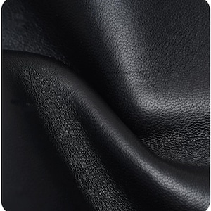 May include: A close-up view of black leather material featuring a distinct pebbled texture. The surface shows natural folds and soft curves, highlighting the matte finish and the irregular grain pattern characteristic of the hide's organic surface.