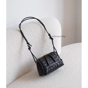 K&ouml;nnte beinhalten: Eine schwarze Crossbody-Tasche aus geflochtenem Leder mit goldenem Rei&szlig;verschluss und einem langen, d&uuml;nnen Riemen. Die Tasche ist rechteckig und weist ein detailliertes Webmuster auf. Der Riemen ist oben geknotet.