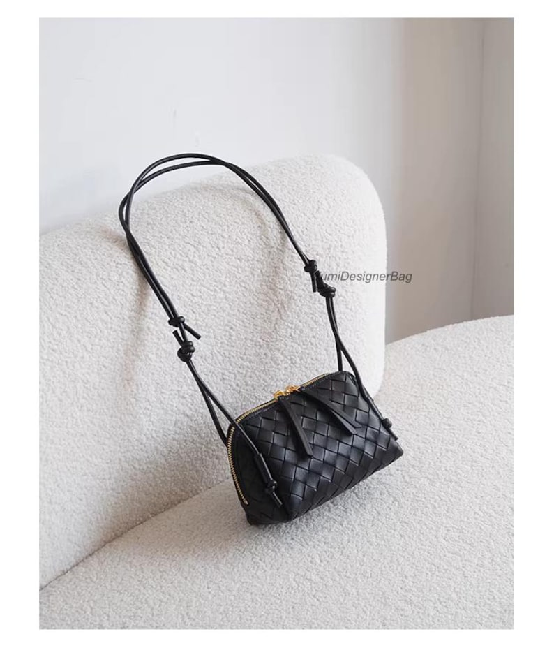 K&ouml;nnte beinhalten: Eine schwarze Crossbody-Tasche aus geflochtenem Leder mit goldenem Rei&szlig;verschluss und einem langen, d&uuml;nnen Riemen. Die Tasche ist rechteckig und weist ein detailliertes Webmuster auf. Der Riemen ist oben geknotet.