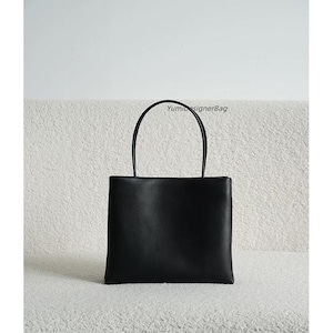 May include: A black rectangular handbag with a smooth finish and two thin, rounded top handles. The bag features a clean, structured design with a flat base and the text YumiDesignerBag printed in small black letters near the top right handle.
