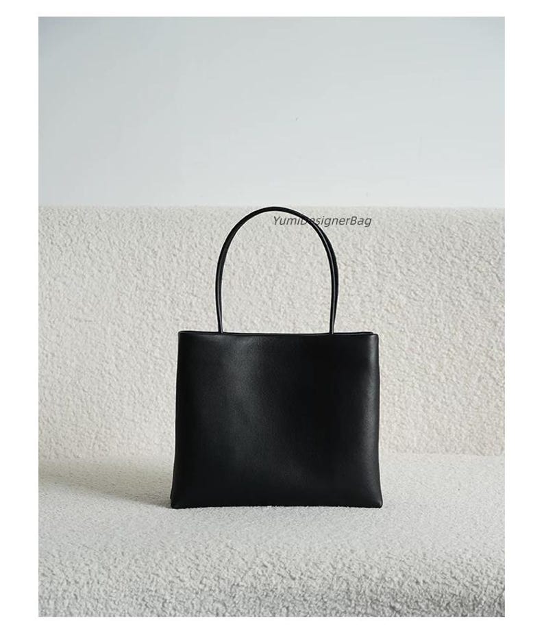 May include: A black rectangular handbag with a smooth finish and two thin, rounded top handles. The bag features a clean, structured design with a flat base and the text YumiDesignerBag printed in small black letters near the top right handle.