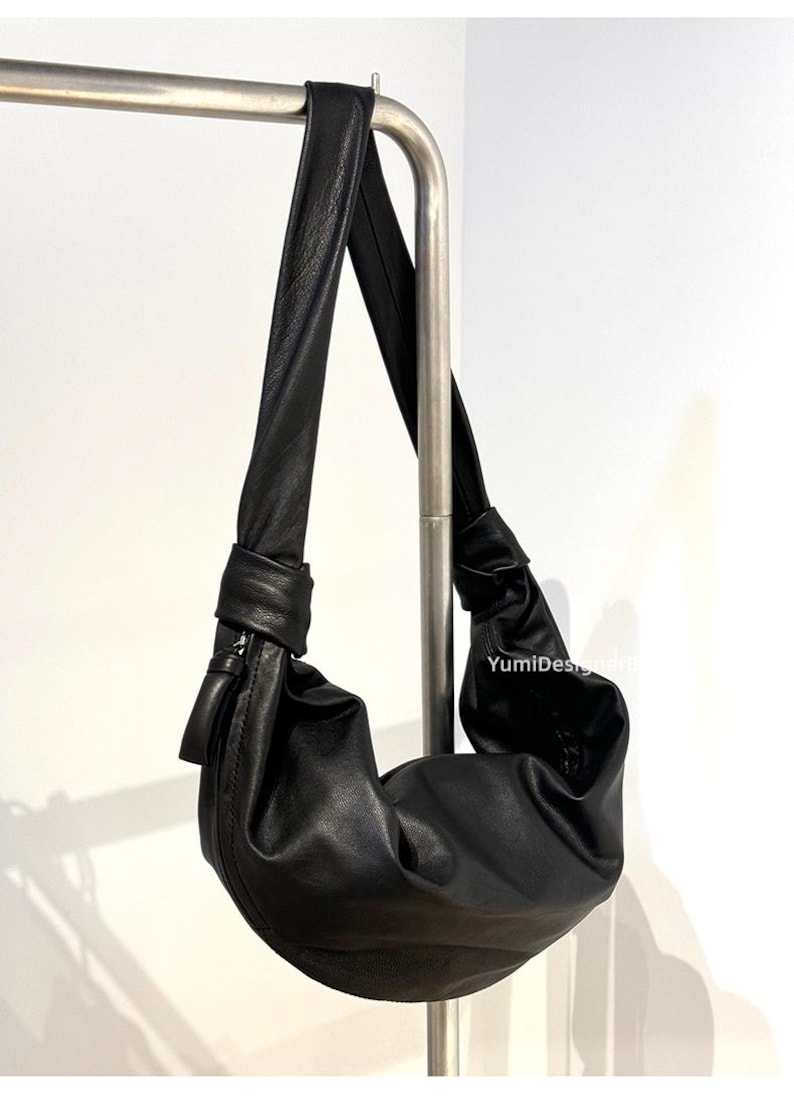 May include: A black leather shoulder bag with a knotted strap and a curved, crescent-shaped body. The bag hangs from a silver metal bar against a white background. The bag has a zipper closure and a small tassel.