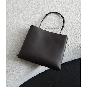 May include: A dark brown rectangular handbag with a smooth finish and two thin, rounded top handles. The bag features the text YumiDesignerBag printed in small, dark letters near the top center, providing a clean and structured appearance.