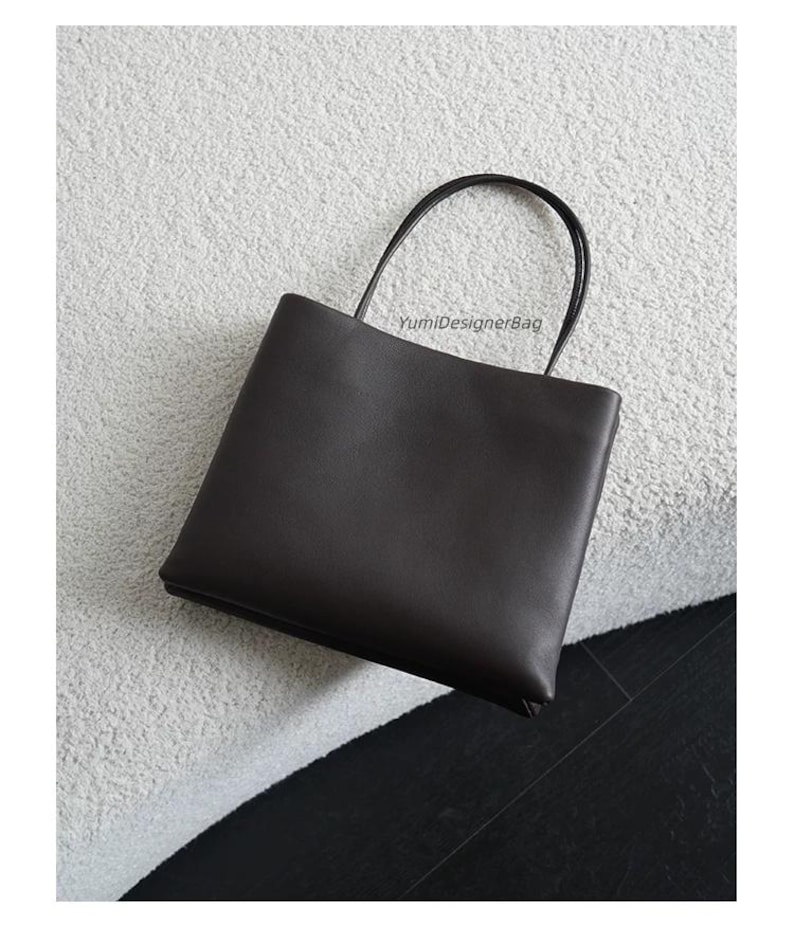 May include: A dark brown rectangular handbag with a smooth finish and two thin, rounded top handles. The bag features the text YumiDesignerBag printed in small, dark letters near the top center, providing a clean and structured appearance.