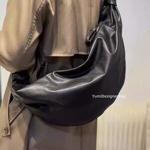 May include: A black leather shoulder bag with a knotted strap. The bag is crescent-shaped and appears to be made of soft leather. The text "YumiDesignerBag" is visible on the bag.