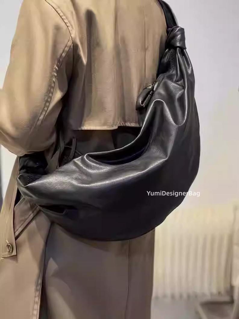 May include: A black leather shoulder bag with a knotted strap. The bag is crescent-shaped and appears to be made of soft leather. The text "YumiDesignerBag" is visible on the bag.