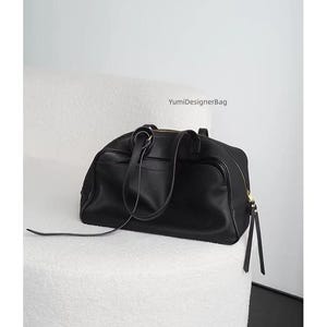 May include: A black leather duffel bag with a top handle and a shoulder strap. The bag has a gold zipper and is sitting on a white, textured surface. The text "YumiDesignerBag" is visible.