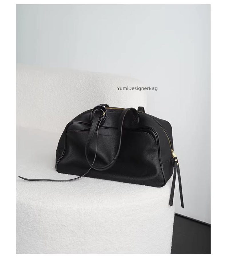 May include: A black leather duffel bag with a top handle and a shoulder strap. The bag has a gold zipper and is sitting on a white, textured surface. The text "YumiDesignerBag" is visible.